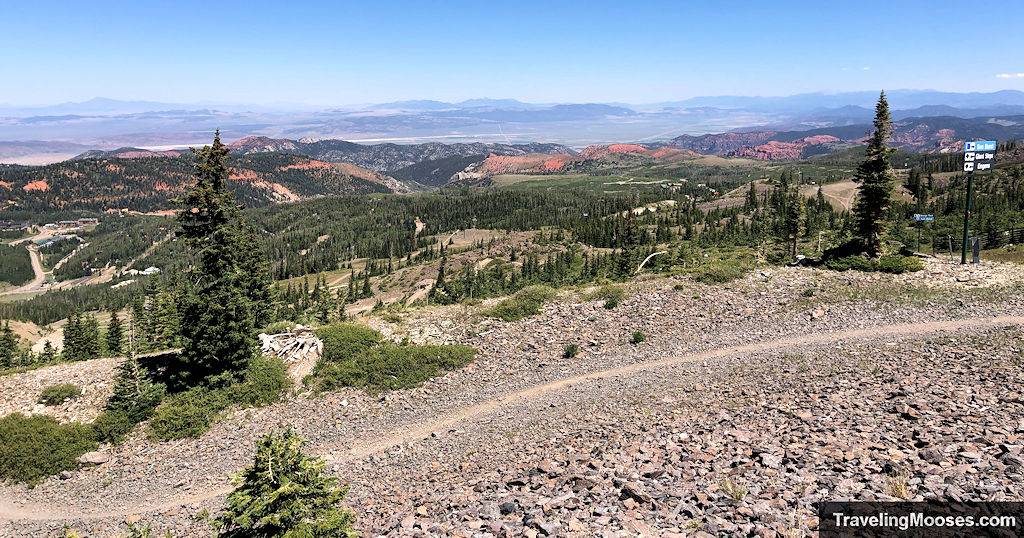 View from the Color Flow trail at Brian Head showing rocky singletrack, forested slopes, and red cliffs in the distance on a clear summer day.